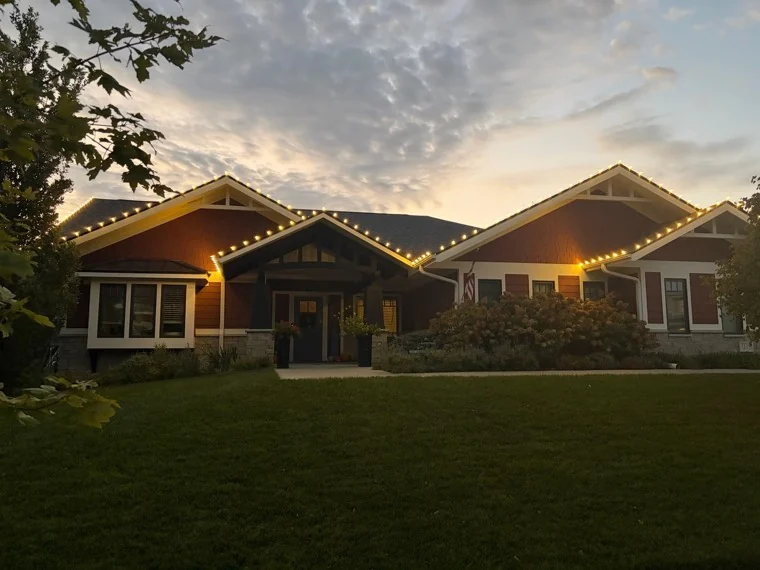 Warm white Christmas lights traced along every gable and roofline of a craftsman home at dusk in Fallbrook, Lincoln, NE